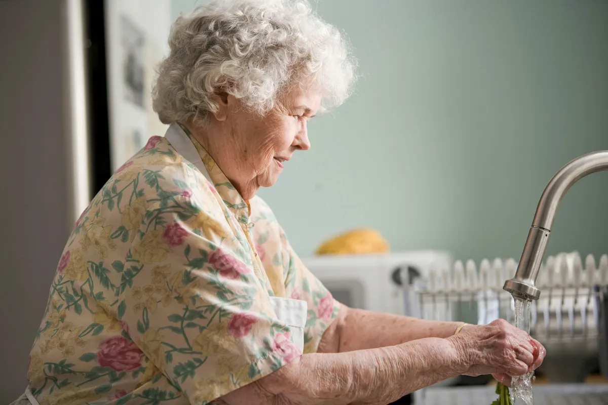 an elderly woman at a care home washing vegetables in the sink